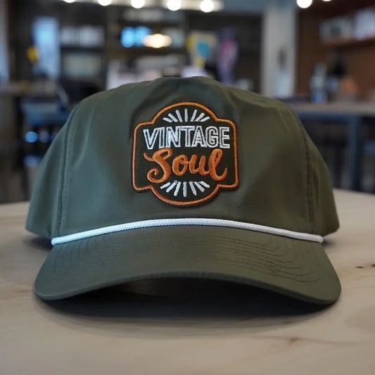 Olive green snapback hat with Vintage Soul embroidered patch in orange and white, rope trim detail, displayed on a wood surface